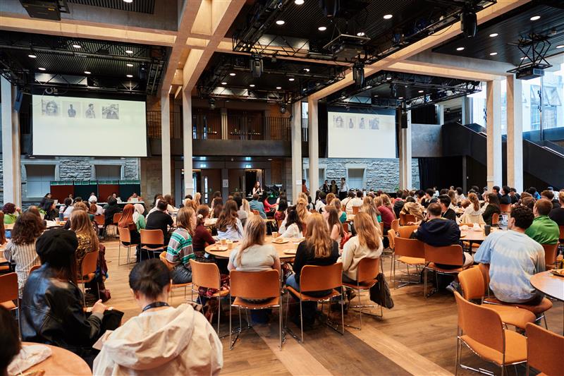 A large group of people sit at round tables in a spacious conference hall, listening to a speaker on stage. Presentation slides with photos are projected on screens at the front of the room.