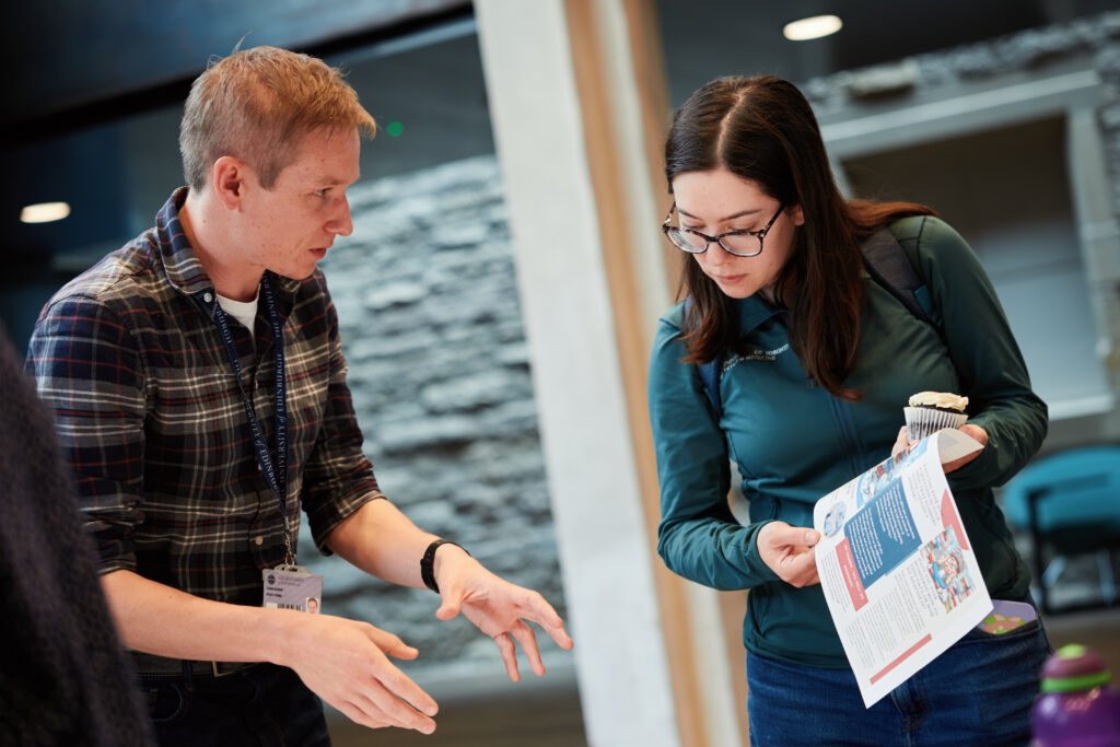 A man explains something to a woman holding an open magazine as they stand indoors. The woman looks at the magazine while the man gestures with his hands. Both appear engaged in conversation.