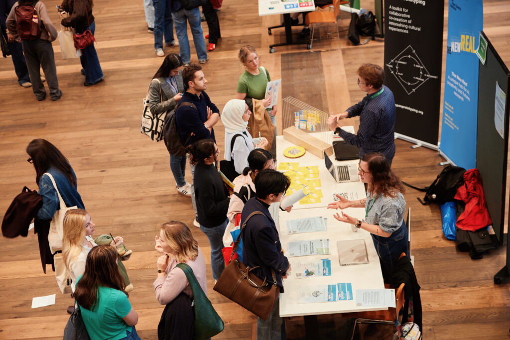 A group of people interact at tables during an indoor event or fair. Some are standing in line, while others engage with booth representatives. Posters, flyers, and informational materials are laid out on the tables.