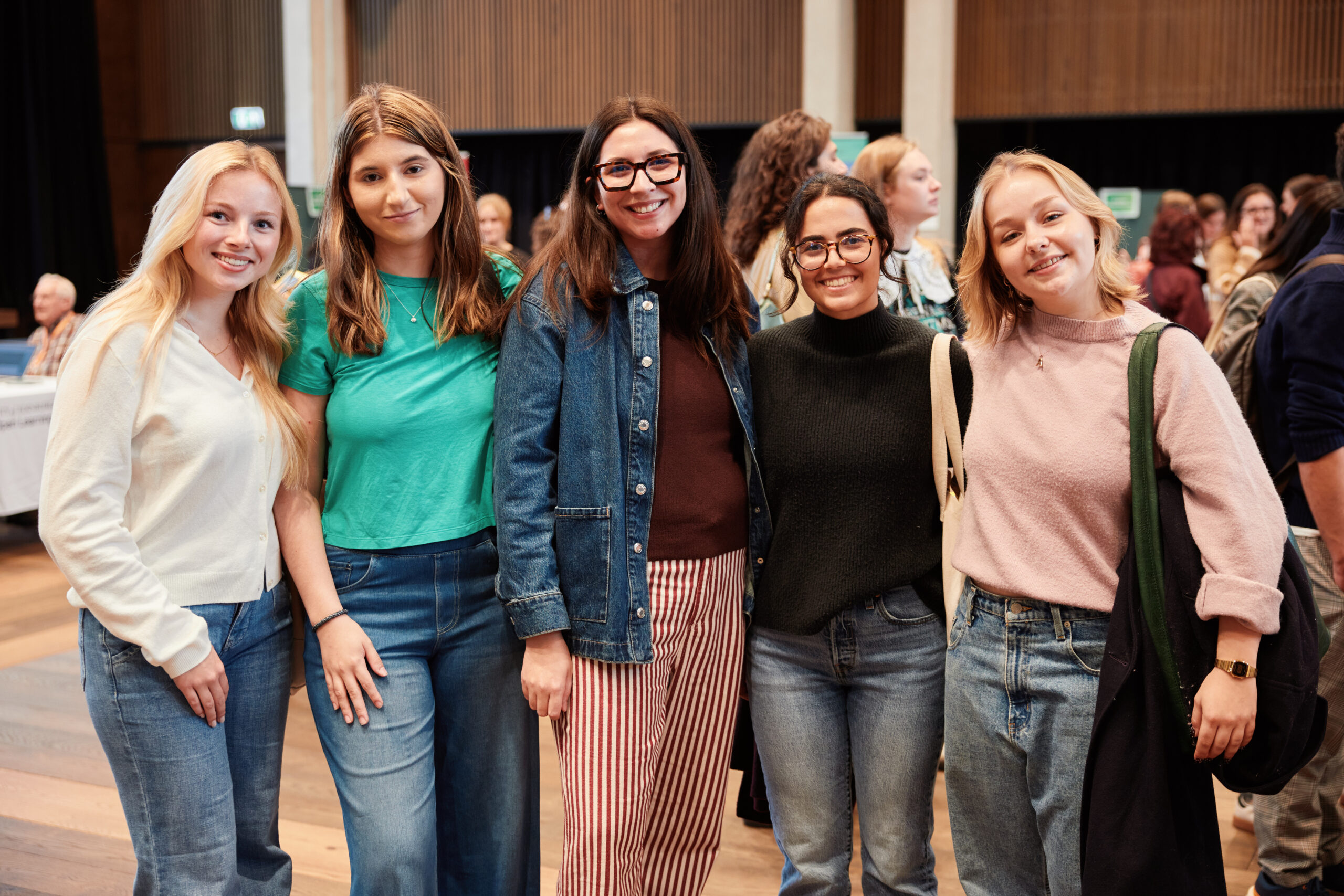Five women stand together indoors, smiling at the camera. They are casually dressed in jeans and sweaters or jackets. Other people are visible in the background, suggesting a social or community event.