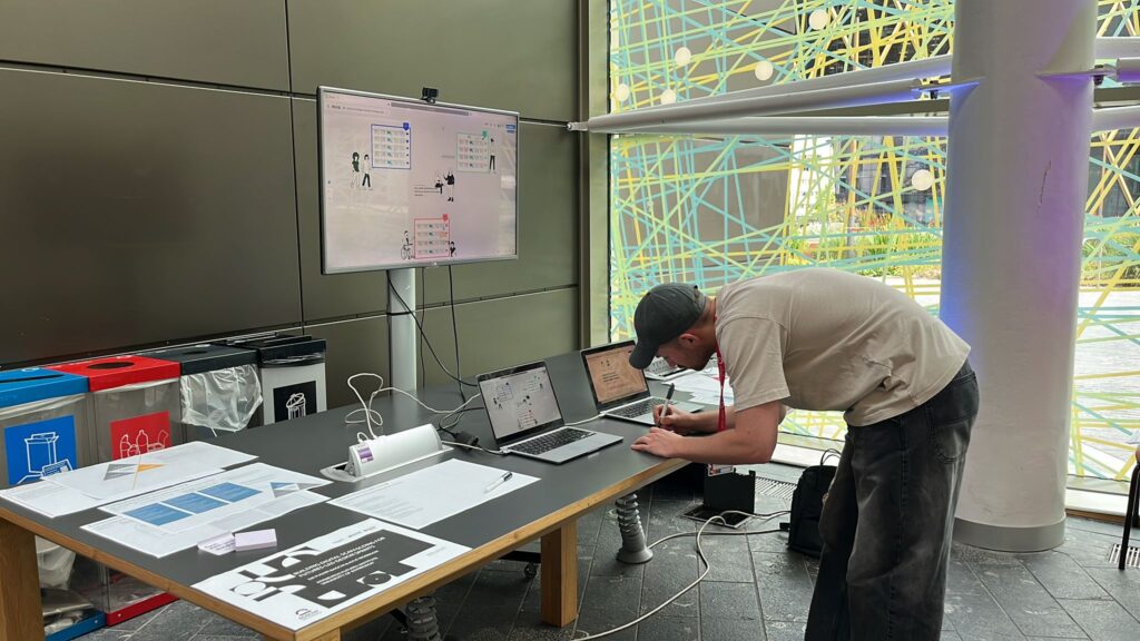 A person leans over a table with papers, two open laptops, and a monitor displaying diagrams. The table is set up near large windows with colorful lines and circles on the glass. Recycling bins are visible on the left.