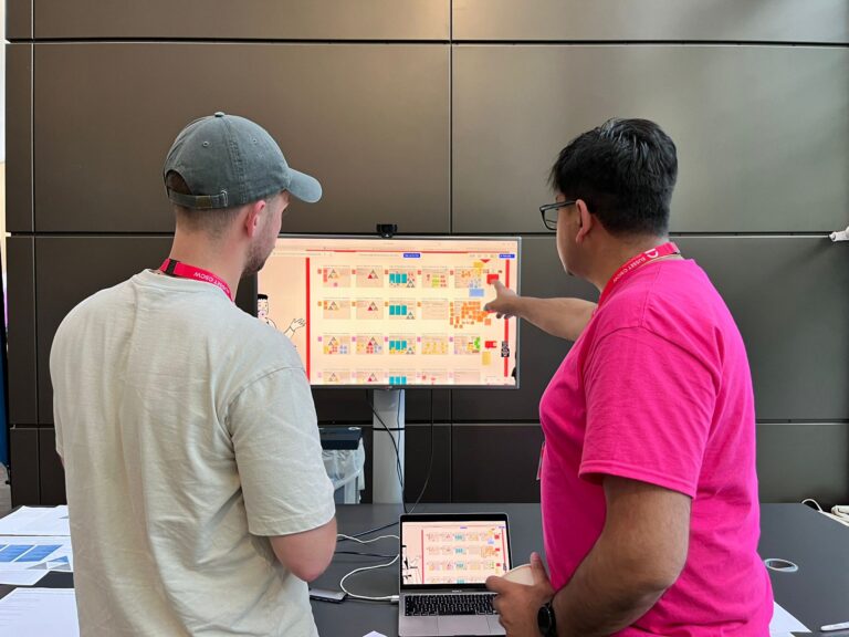 Two people stand near a desk, looking at a large monitor with colorful charts and diagrams. One person in a pink shirt points at the screen while the other listens. A laptop showing similar content is on the desk.