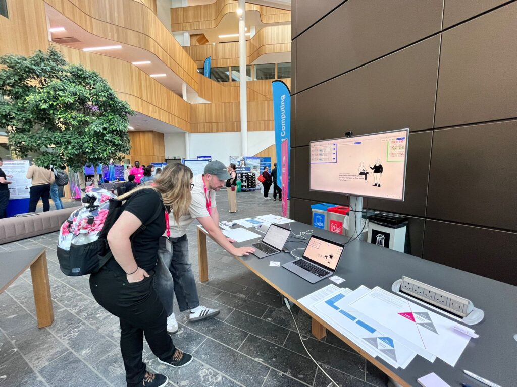 Two people stand at a table with laptops, papers, and a large monitor displaying a cartoon in a modern, open indoor space with posters and booths in the background.