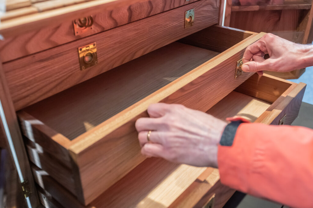 A person wearing an orange sleeve and a gold ring opens a wooden drawer in a wooden cabinet with brass hardware. The drawer is empty, and several compartments are visible.