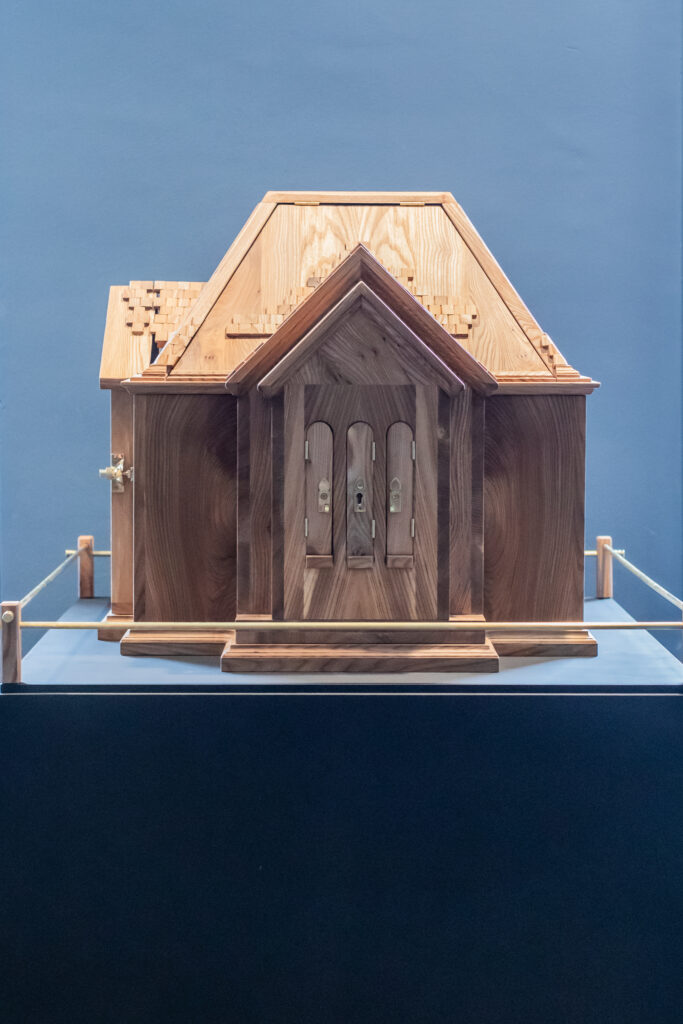A detailed wooden model of a house displayed on a stand, surrounded by a rope barrier, set against a plain blue background.