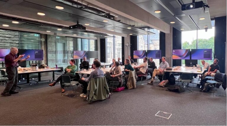 A group of people sit at tables in a modern conference room, listening to a presenter standing at the front. Large windows and multiple monitors are visible in the background.
