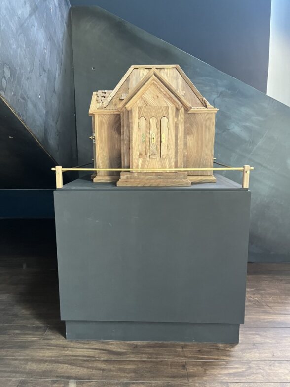 A small, wooden, house-shaped box with intricate carvings is displayed on a dark pedestal, bordered by a thin gold rope barrier. The background features dark, angular walls and wooden flooring.