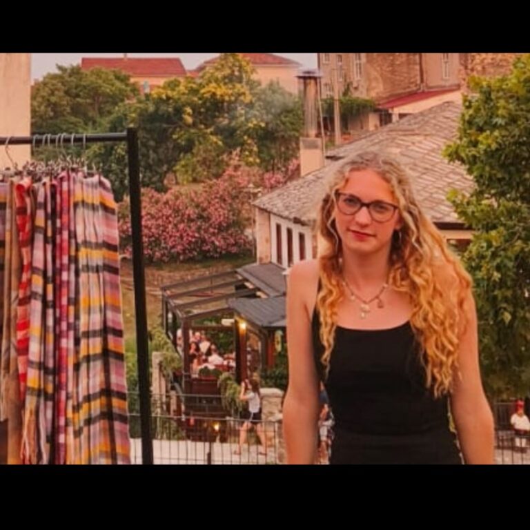 A woman with long curly blonde hair and glasses stands next to a rack of colorful scarves outdoors, with trees, flowers, and buildings in the background. She is wearing a black sleeveless top.