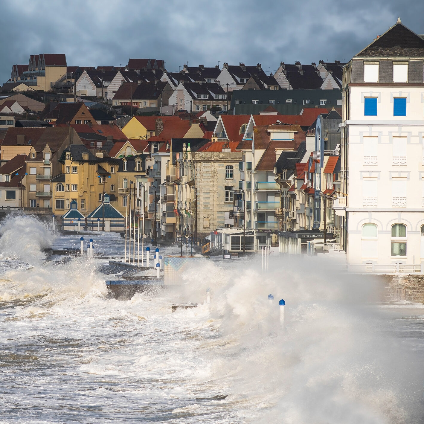 Large waves crash against a seaside town’s waterfront, with spray rising high. Colorful houses and buildings line the shore under a cloudy sky, suggesting a stormy or windy day.
