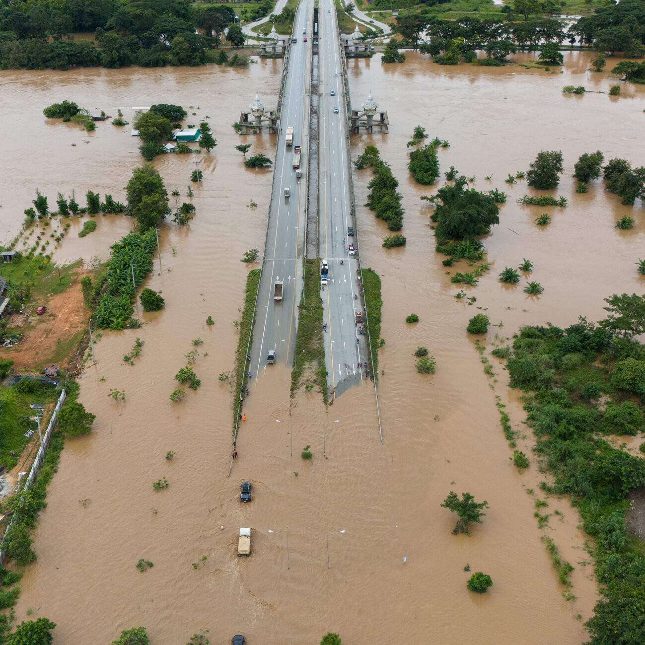 Aerial view of a flooded area with a highway running through the center; vehicles drive cautiously on the partially submerged road, and water covers the surrounding land and buildings.