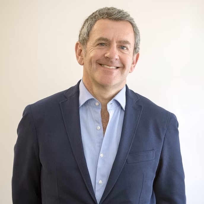 A middle-aged man with short gray hair smiles while standing against a plain light background. He is wearing a blue blazer over a light blue dress shirt, which is unbuttoned at the top.