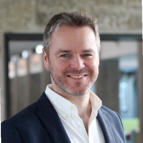 A man with short, grayish hair and a trimmed beard, wearing a white collared shirt and dark blazer, smiles while standing indoors in front of a blurred background with stone and glass elements.