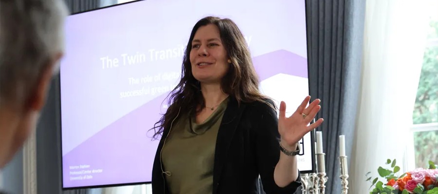 A woman with long brown hair speaks and gestures with one hand during a presentation. She stands in front of a screen displaying a slide titled "The Twin Transformation" in a bright, modern room.