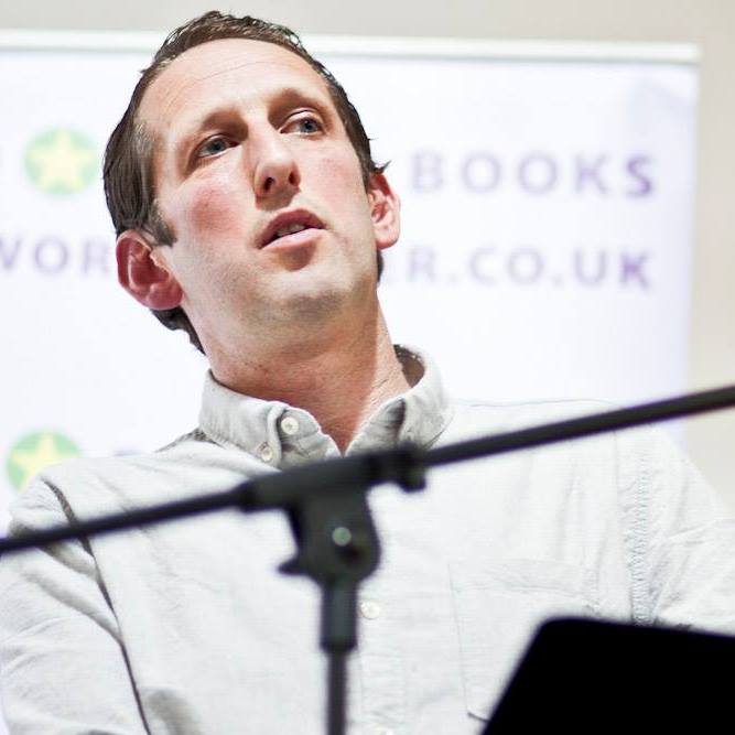 A man in a light-colored button-up shirt speaks in front of a microphone. Behind him is a blurred banner with partial text, including "BOOKS" and a website URL ending in ".CO.UK".