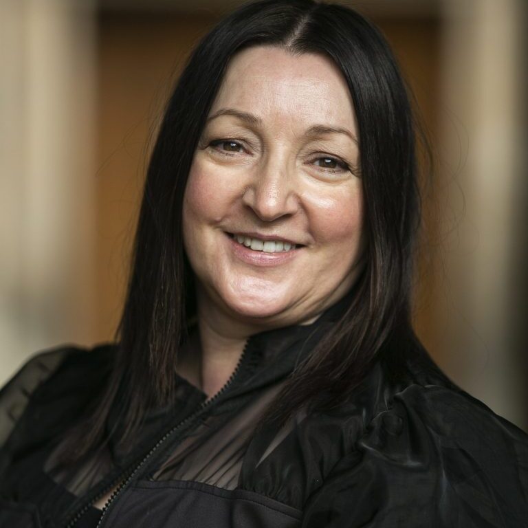 A woman with long dark hair, wearing a black sheer-sleeved top, smiles warmly at the camera. The background is softly blurred with warm tones.