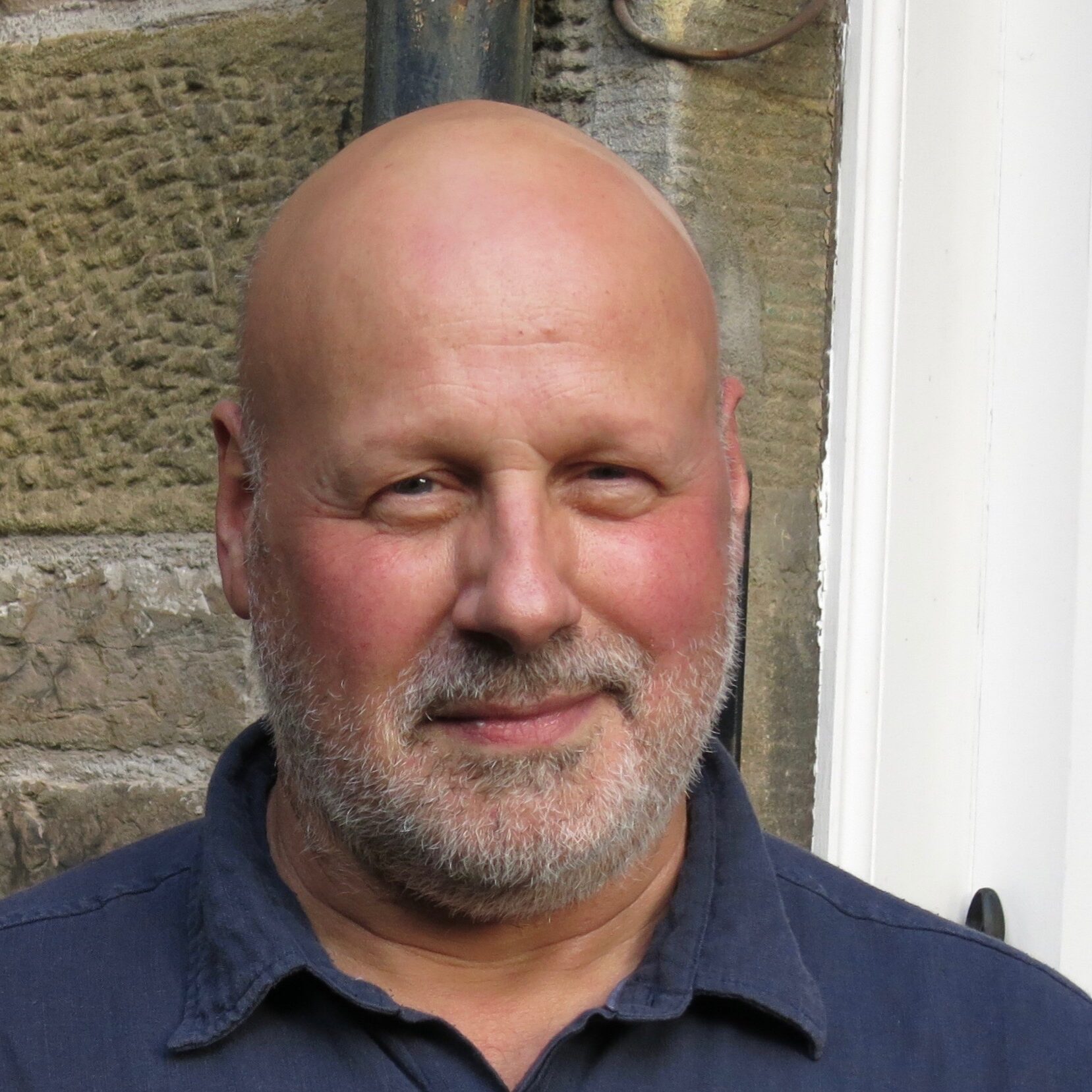 A middle-aged man with a bald head and gray beard, wearing a dark blue shirt, stands in front of a stone wall and white window frame, smiling gently at the camera.