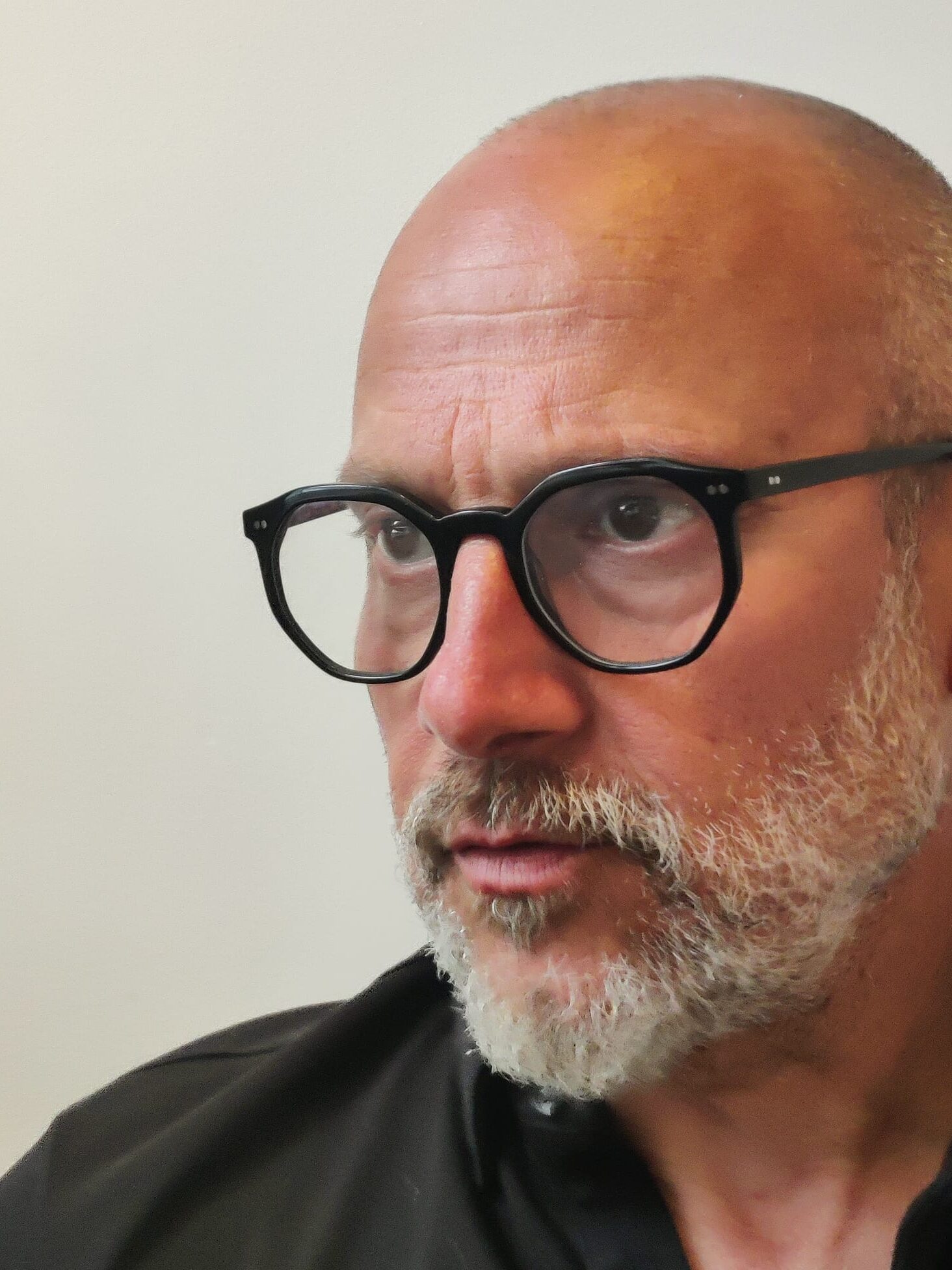 A middle-aged man with a shaved head, gray beard, and black-framed glasses looks thoughtfully to the left. He is wearing a black shirt and is set against a plain, light-colored background.