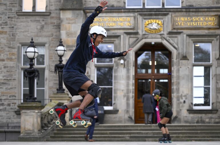 A person wearing a helmet and knee pads jumps on roller skates in front of a stone building, while another person on skates moves in the background. The building has large windows and signs with gold lettering above the entrance.