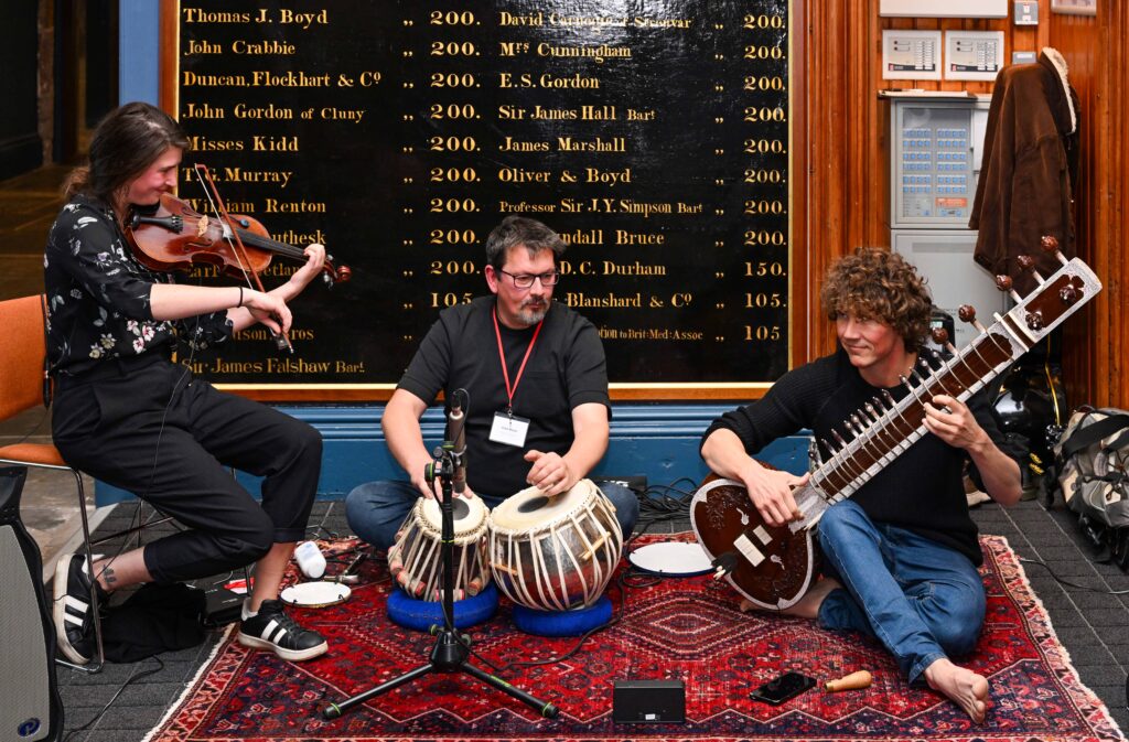 Three musicians perform together indoors: one playing a violin, one playing tabla drums, and one playing a sitar. They are seated on a patterned rug in front of a wall with lists of names.