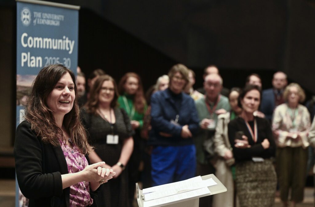 A woman speaks at a podium while a group of people stand and listen in the background. A banner reads "The University of Edinburgh Community Plan 2018-30." The atmosphere appears to be that of a formal event or presentation.