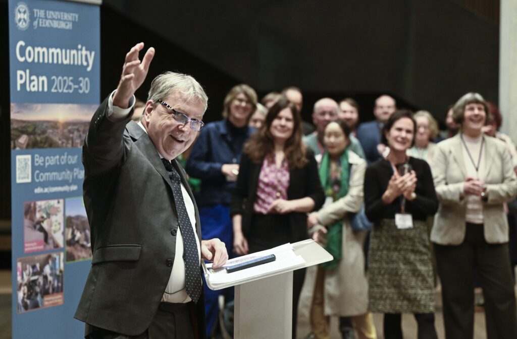 A man in a suit stands at a podium, smiling and waving to an applauding audience. Behind him is a group of people and a sign that reads "Community Plan 2025-30, The University of Edinburgh.