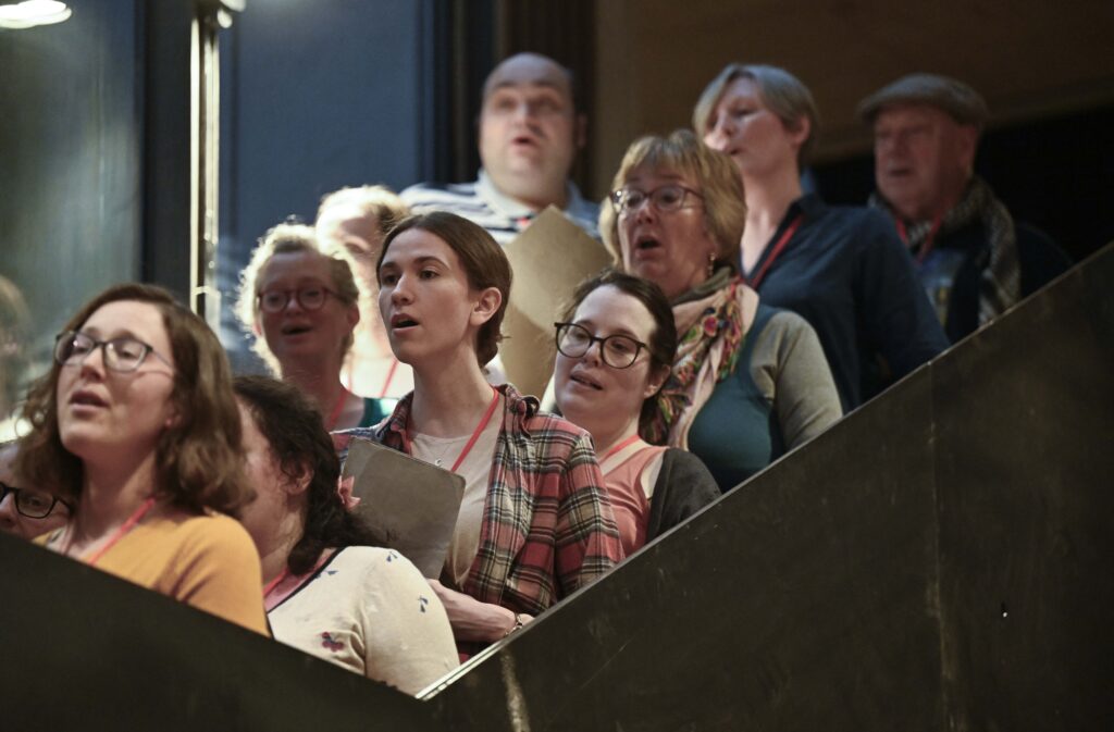 A group of people, both men and women, stand on a staircase singing together, each holding sheet music. They appear to be in a choir rehearsal, focused on their performance.
