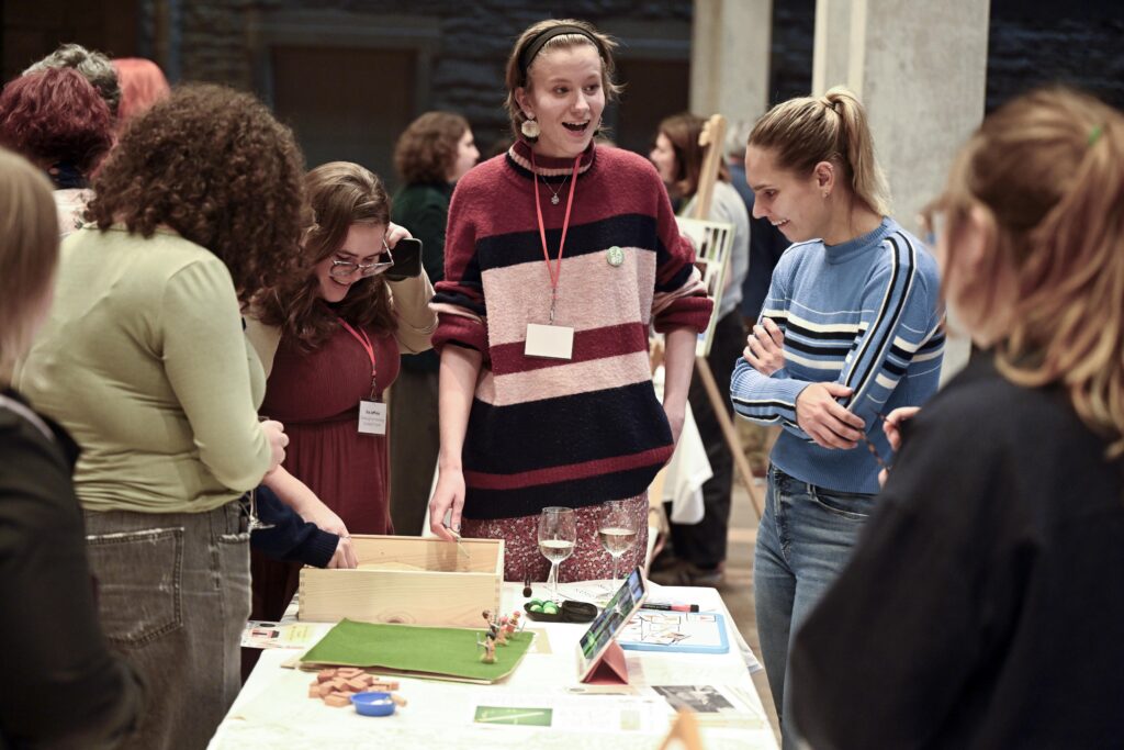 A group of people gather around a table with games and papers, chatting and smiling in a bright indoor space. One woman in a striped sweater stands and talks while others look at the items on the table.