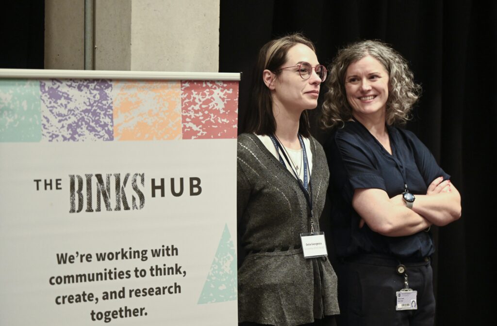 Two women stand next to a sign that reads “The Binks Hub: We’re working with communities to think, create, and research together.” One woman is wearing glasses; both have name badges and appear to be at an event.