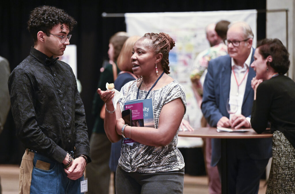 A woman holding a booklet and a snack talks to a man at a professional networking event, while other people converse in the background around high tables.