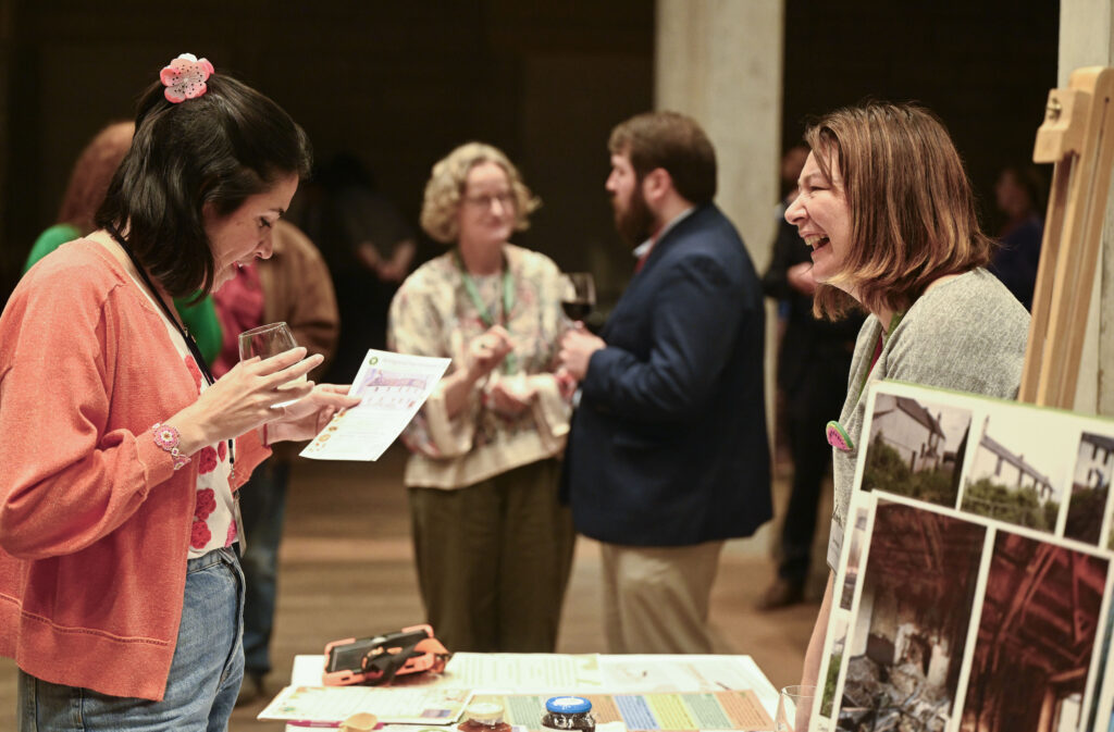 Two women engage in a lively conversation at an indoor event table, smiling and holding papers. Other people socialize in the background, drinks in hand. Various informational materials and photos are displayed on the table.