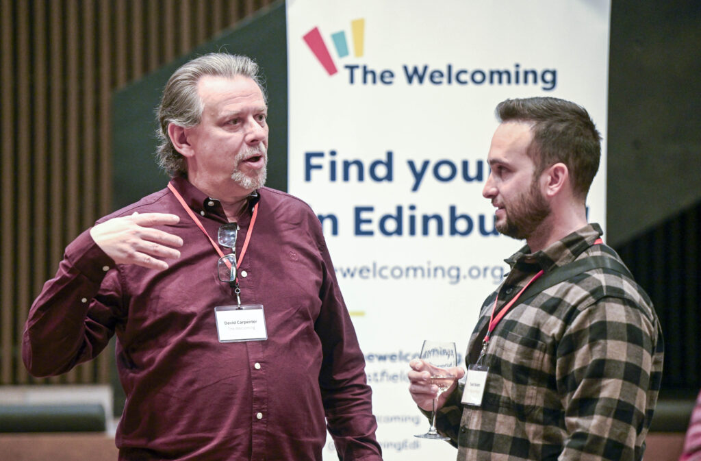 Two men wearing name tags converse at an event, with one holding a drink. A banner behind them reads "The Welcoming" and "Find your in Edinburgh," promoting thewelcoming.org.uk.