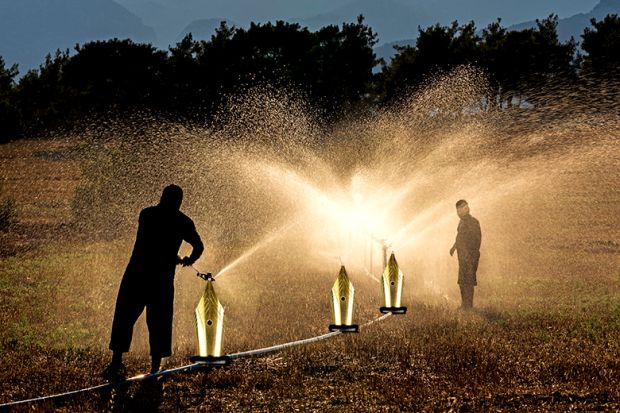 Two people stand in a sunlit field, using hoses to spray water. The water creates a misty effect, and in the foreground, large fountain pen nibs are edited onto the hose nozzles, resembling fountains. Trees silhouette the background.