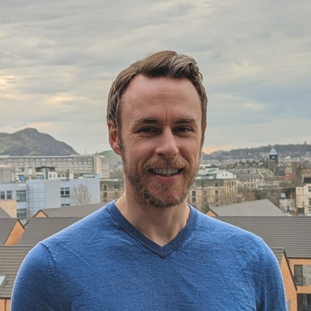 A man with short brown hair and a beard, wearing a blue sweater, stands outdoors smiling with buildings, rooftops, and distant hills under a cloudy sky in the background.