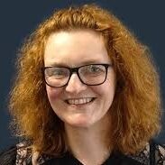 A person with curly red hair, glasses, and a black top is smiling in front of a dark blue background.