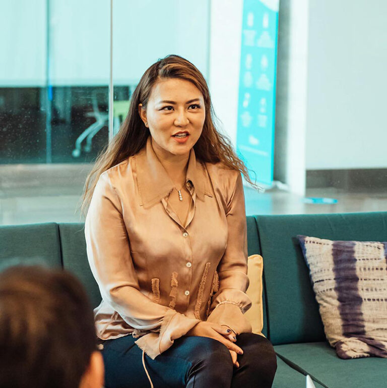 A woman with long brown hair, wearing a shiny beige blouse, sits on a teal couch speaking to others. There is a pillow beside her and a glass wall in the background.