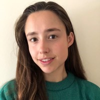 A young woman with long brown hair and brown eyes, wearing a green sweater, smiles softly while standing in front of a plain light-colored wall.