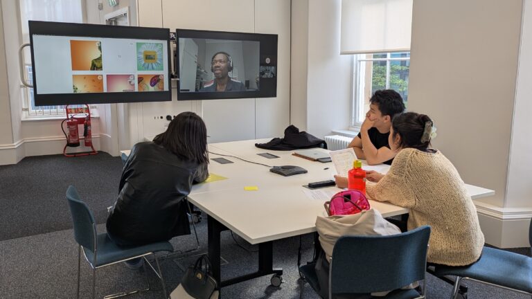 Three people sit around a table in a meeting room, watching a video call on two large screens. The screens display a speaker and images. Papers, laptops, and water bottles are on the table.