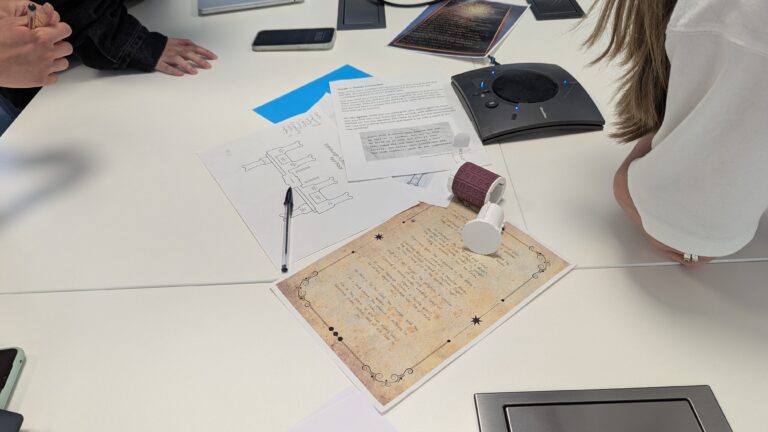 Three people gather around a white table with scattered documents, including a vintage-looking paper, diagrams, pens, smartphones, and a speakerphone, suggesting a collaborative work or mystery-solving session.
