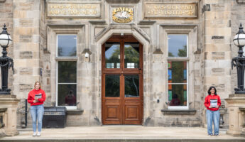 Two people in red shirts stand apart on the steps of a stone building with large wooden doors and gold plaques reading “I was a stranger and ye took me in” and “I was sick and ye visited me.”.