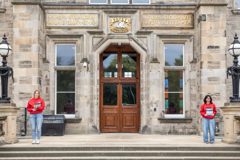 Two people in red shirts stand apart on the steps of a stone building with large wooden doors and gold plaques reading “I was a stranger and ye took me in” and “I was sick and ye visited me.”.