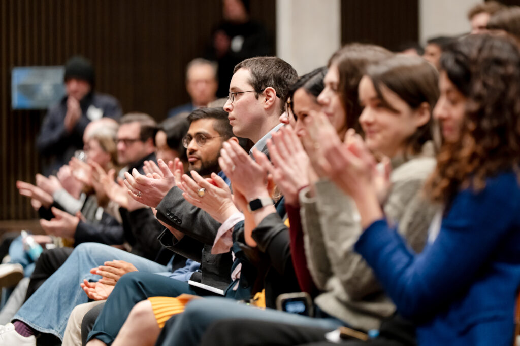 A group of people sitting in rows, attentively clapping and facing forward at an indoor event or presentation. The background is slightly blurred, emphasizing the engaged audience members.