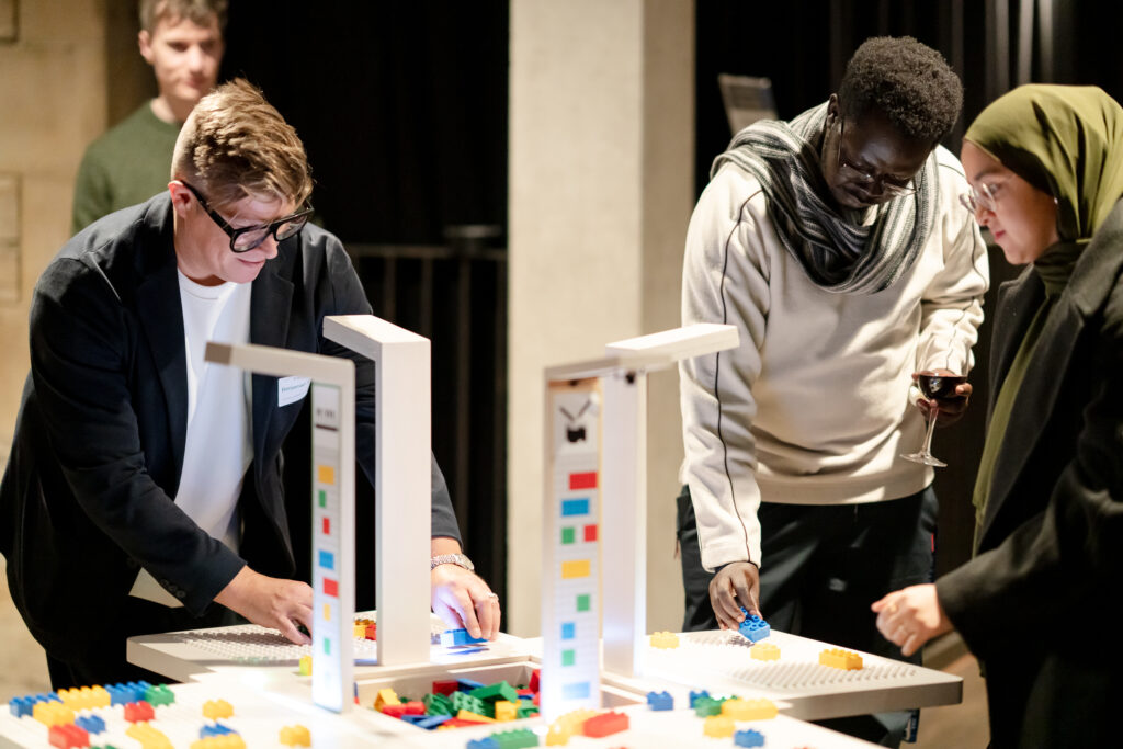 Three people stand around a table building with colorful plastic bricks. One person wears a suit and glasses, another holds a wine glass, and the third wears a headscarf. A fourth person stands in the background, watching.