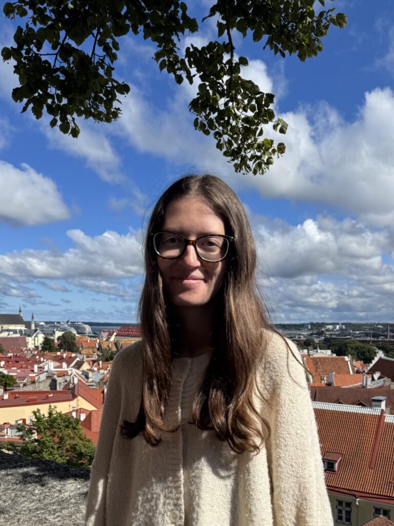 A young person with long brown hair and glasses stands outdoors in sunlight, smiling. Behind them are red-roofed buildings and a blue sky with white clouds. Green tree branches frame the top of the image.