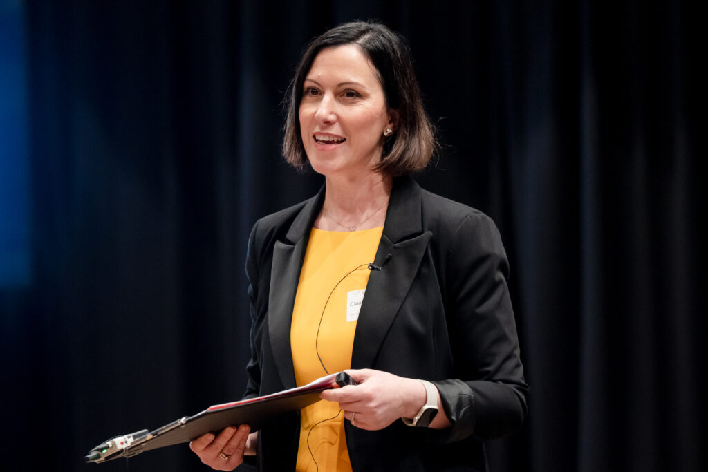 A woman with short dark hair wearing a yellow top and black blazer holds a clipboard and speaks on stage. She has a microphone attached to her shirt and stands in front of a dark, blurred background.