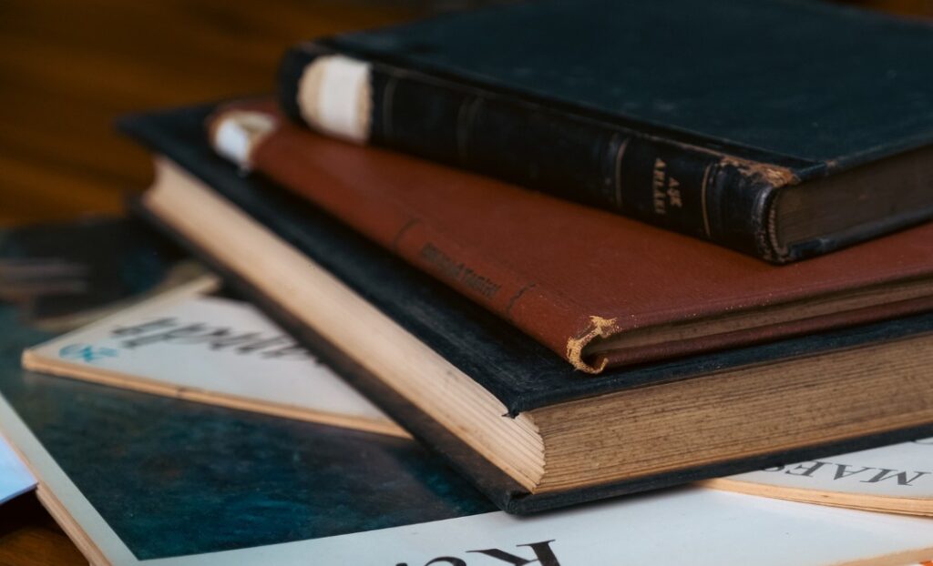 A close-up of a stack of old books and magazines on a wooden surface, with worn covers and frayed edges, suggesting they have been well-used and read many times.