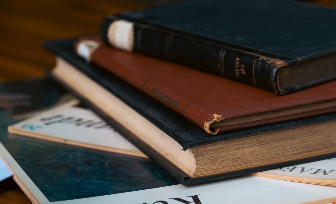 A close-up of a stack of old books and magazines on a wooden surface, with worn covers and frayed edges, suggesting they have been well-used and read many times.