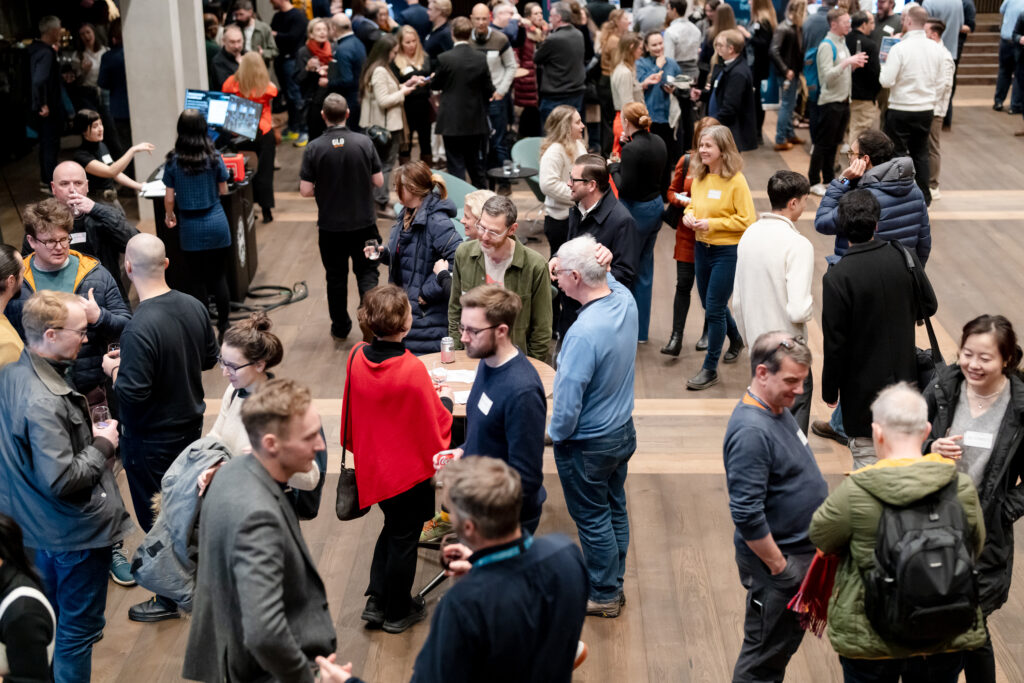 A large group of people mingle and talk at an indoor event. Most are standing in small groups, some holding drinks, and many wear name tags. The atmosphere appears lively and social, with diverse attendees.