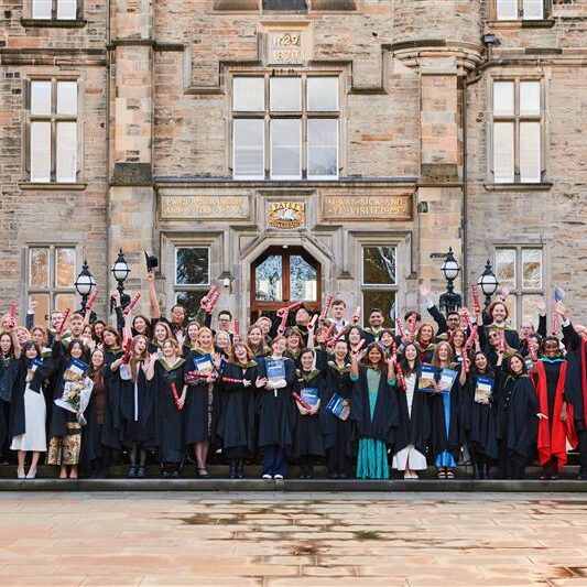 A large group of graduates in caps and gowns stand smiling and celebrating in front of a historic stone building, holding certificates and raising their arms in joy.