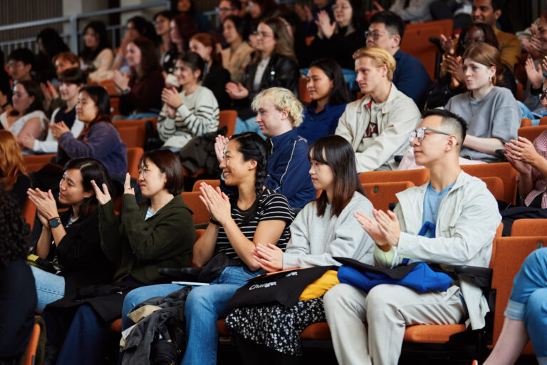A diverse group of people sits in auditorium seats, attentively watching and applauding something on stage. Most individuals are smiling and focused, with some holding notebooks or bags.