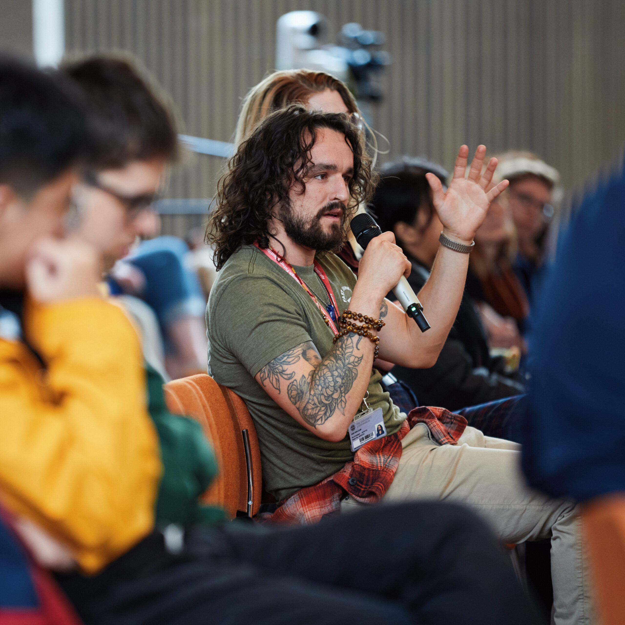 A man with long curly hair and tattoos speaks into a microphone while seated among a group of people in an indoor event. Other attendees listen attentively.